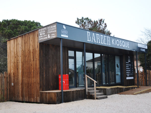 Salon de coiffure-Barber créé à partir d'un conteneur maritime. Bardage en bois brut grisé, coursive couverte et grande vitrine constitue une apparence modernisée de "saloon" de western.