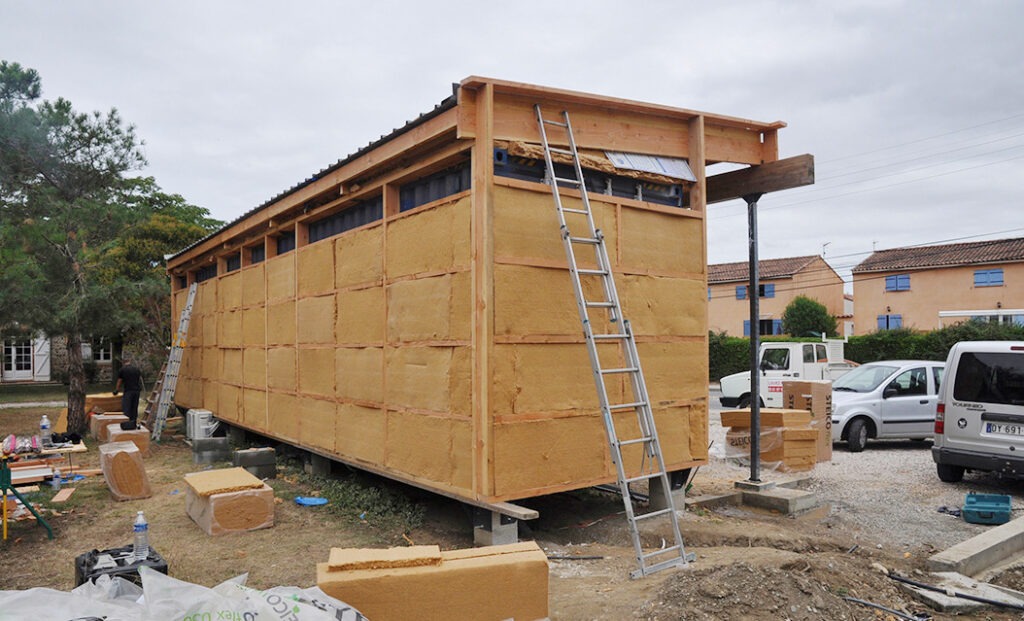Photographie de chantier : Contener maritime habillé d'une ossature secondaire en bois et rempli d'un isolant Le tout supporte un toit rehaussé pour l'isolation et constituant une avancé supportée par une grande poutre en bois sur poteau d'acier.