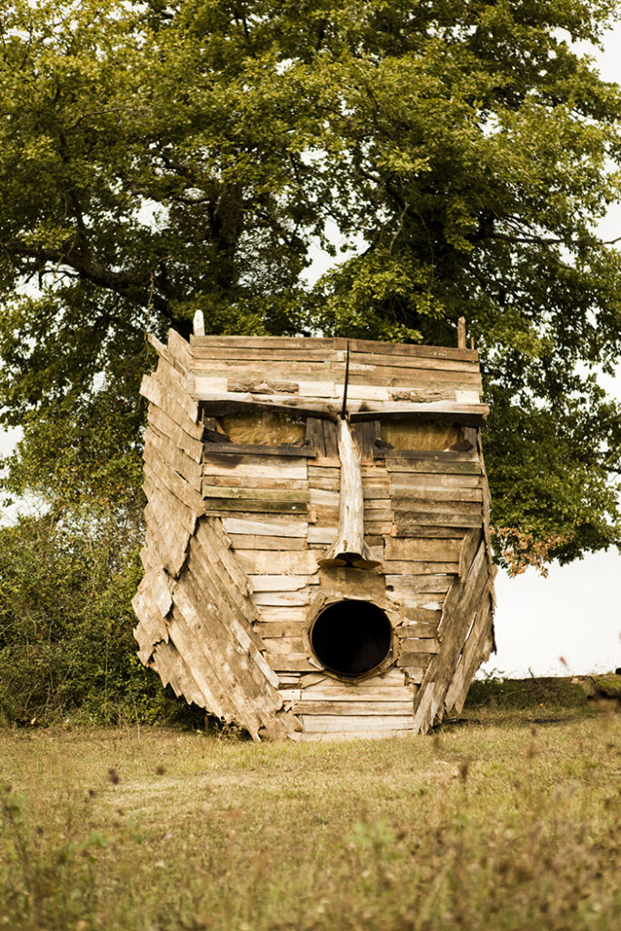 Masque en bois monumental évoquant un Dieu ou un Roi Païen, avec la bouche ouverte (semblant chanter) installé devant un arbre.