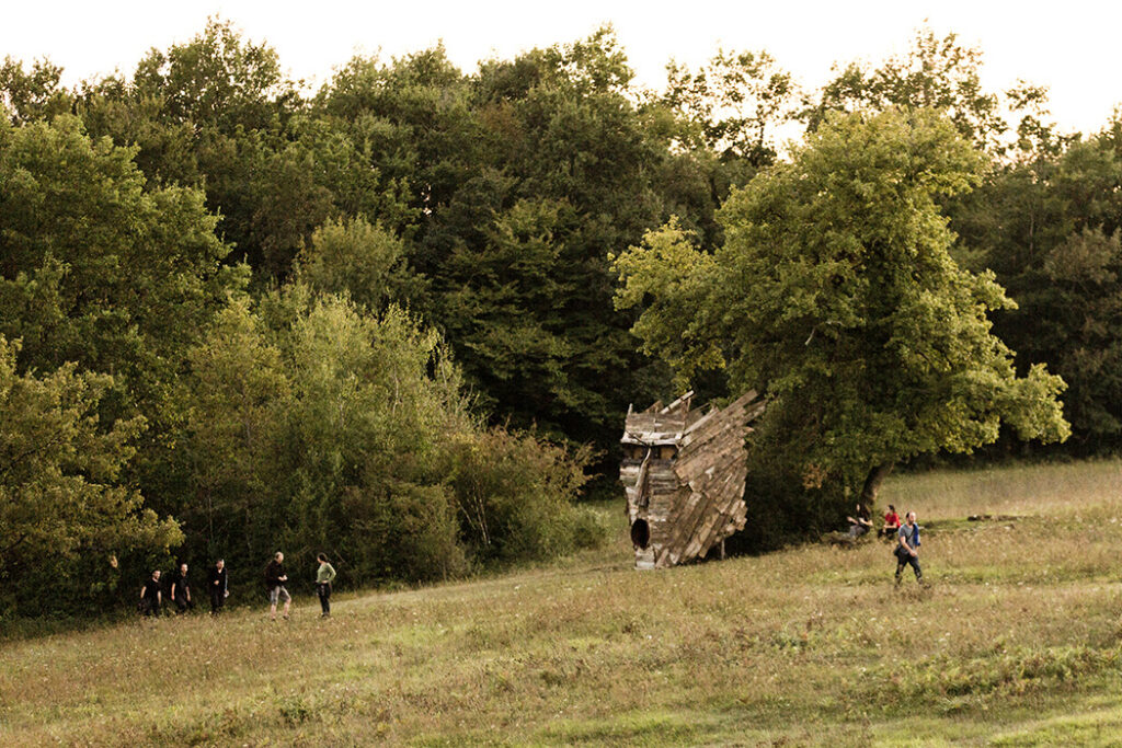 Masque en bois monumental évoquant un Dieu ou un Roi Païen, installé devant un arbre. Festivaliers arrivants sur le site et marchant à proximité.