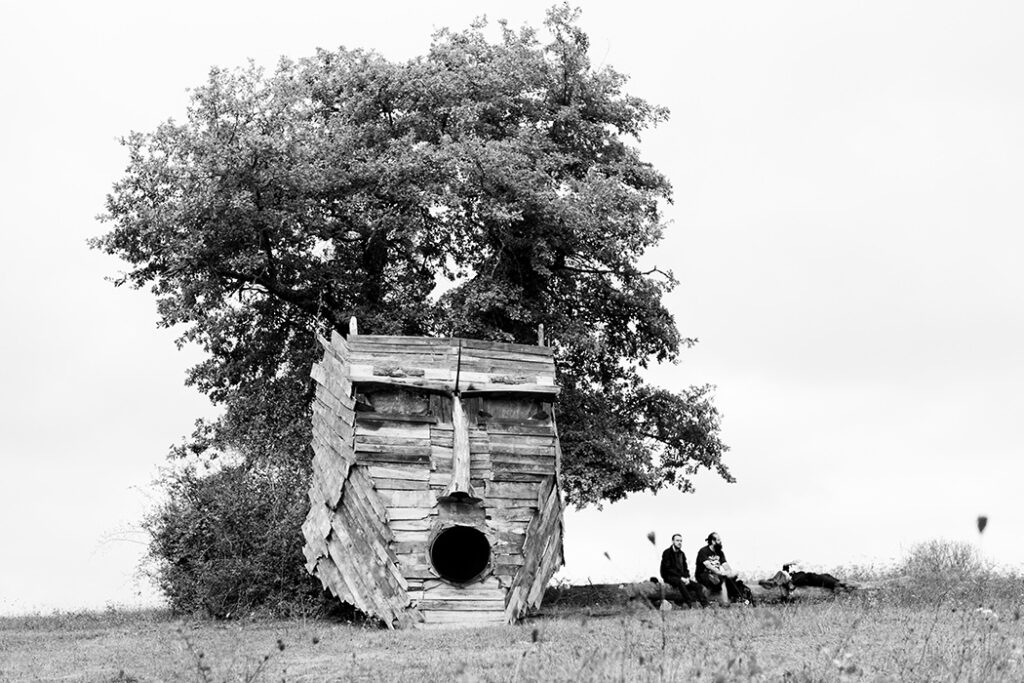 Masque en bois monumental évoquant un Dieu ou un Roi Païen, avec la bouche ouverte (semblant chanter) installé devant un arbre. Deux festivaliers assis à proximité. Cliché en noir et blanc.