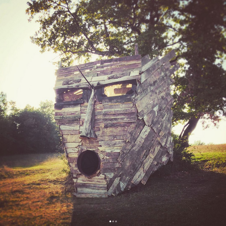 Masque en bois monumental évoquant un Dieu ou un Roi Païen, avec la bouche ouverte (semblant chanter) installé devant un arbre. Prise de vue à contre jour.