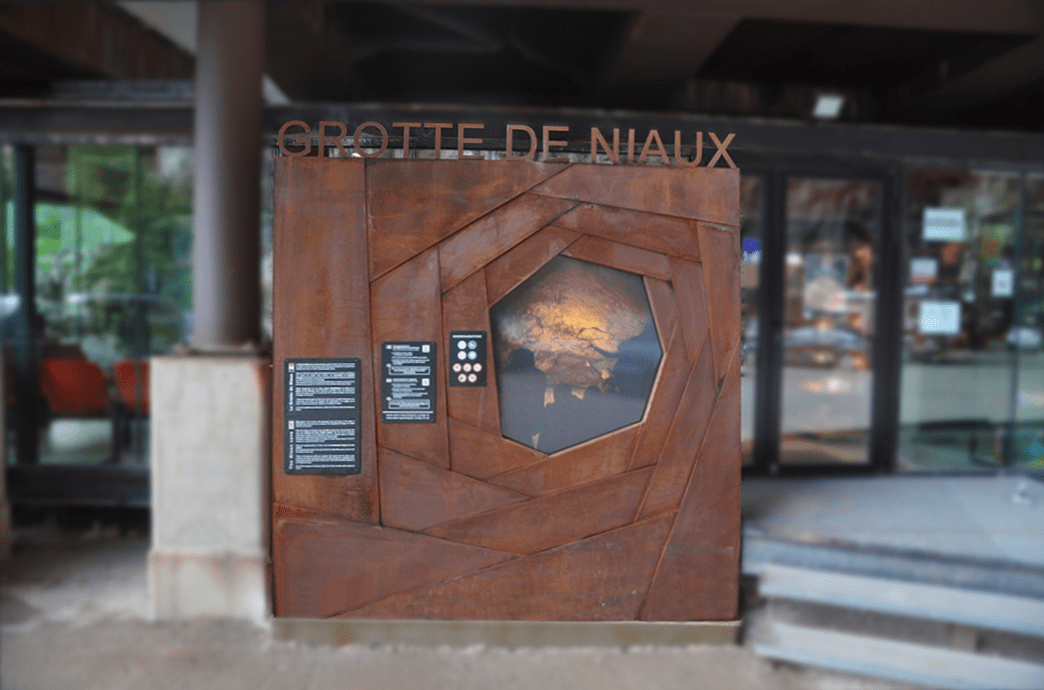 Panneau d'accueil de la grotte de Niaux. c'est une grand carré constitué "d'écailles" en acier Corten dont les formes et l'assemblage symbolisent autant un principe d'obturateur photographique mécanique, qu'une métaphore de la caverne qui s'enfonce vers les profondeurs de la terre