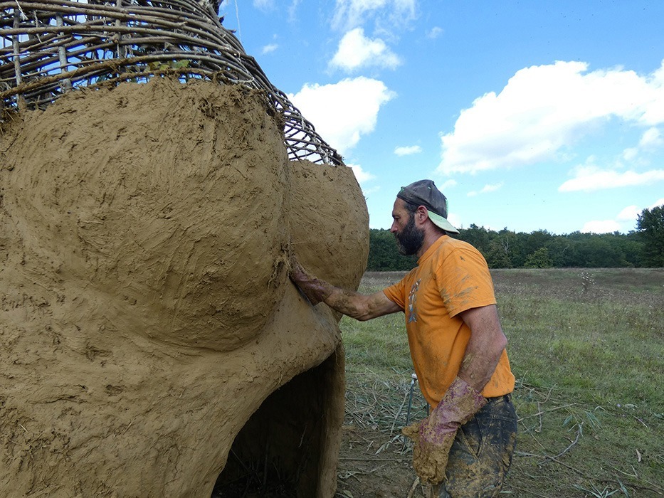 Photographie d'Alexandre Drabzak travaillant à installer le torchis de terre cru sur l'ossature de bois raméal constituant le corps de la Vénus monumentale lors de l'événement musical l'homme sauvage 2024.