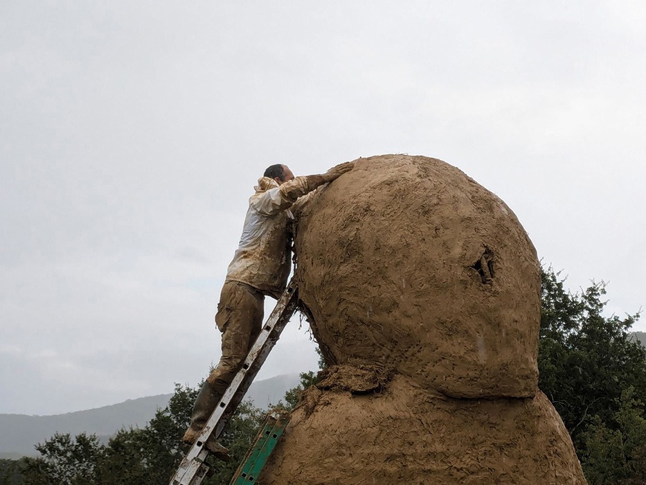 Photographie d'Alexandre Drabzak en haut d'une échelle, travaillant à installer le torchis de terre cru sur la tête de la Vénus monumentale lors de l'événement musical l'homme sauvage 2024.