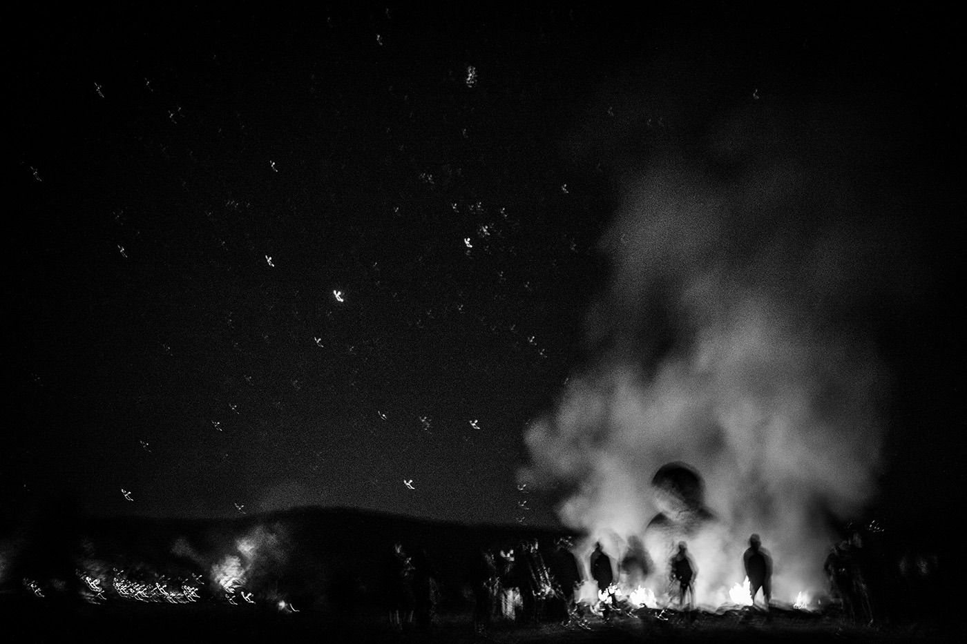 Photographie artistique en noir et blanc montrant les silhouettes du public autour du cercle de feu entourant la Vénus monumentale sur un fond de vaste ciel étoilé.