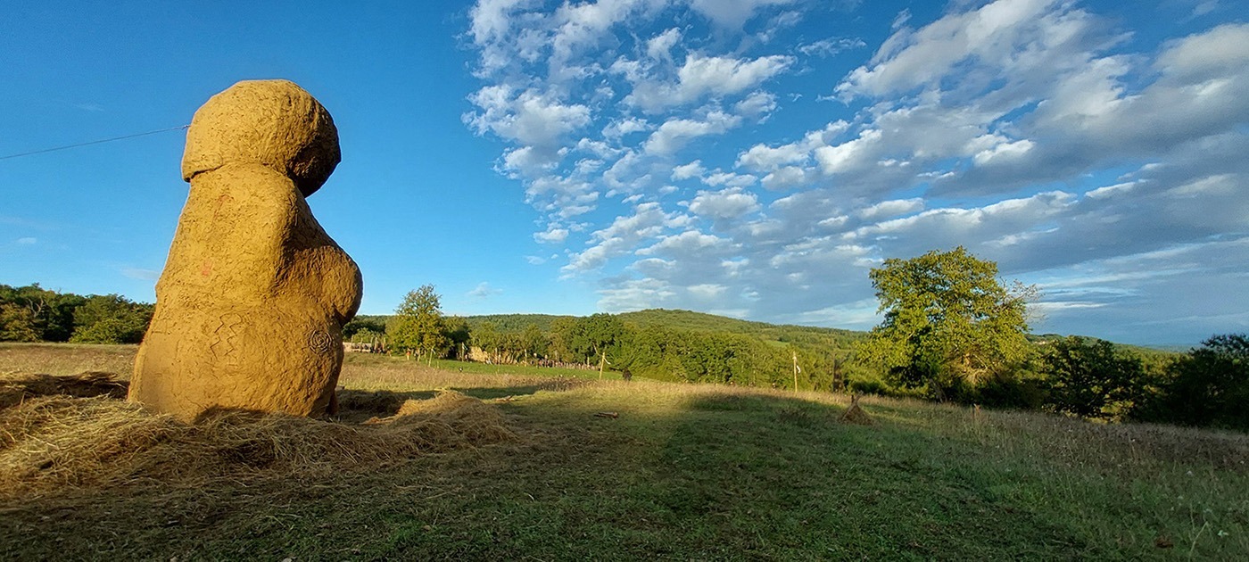 Photographie de la Vénus monumentale en terre cru, dans un décors de prairie,de bois et de ciel bleu avec des trainées de nuages.