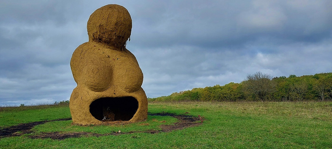 Photographie de la Vénus monumentale en terre cru, dans un décors de prairie et de bois, réalisée à l'occasion de l'événement musical l'homme sauvage 2024.