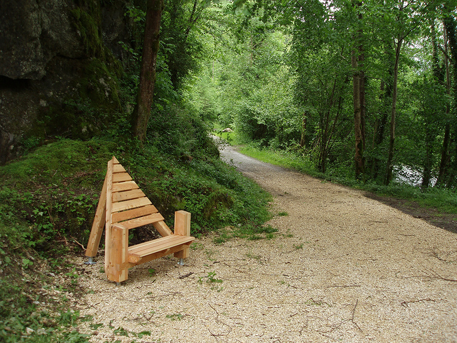 Banc design en bois en forme de triangle et installé au bord d'un chemin de cailloux blanc dans un espace naturel verdoyant et bordé par une rivière.