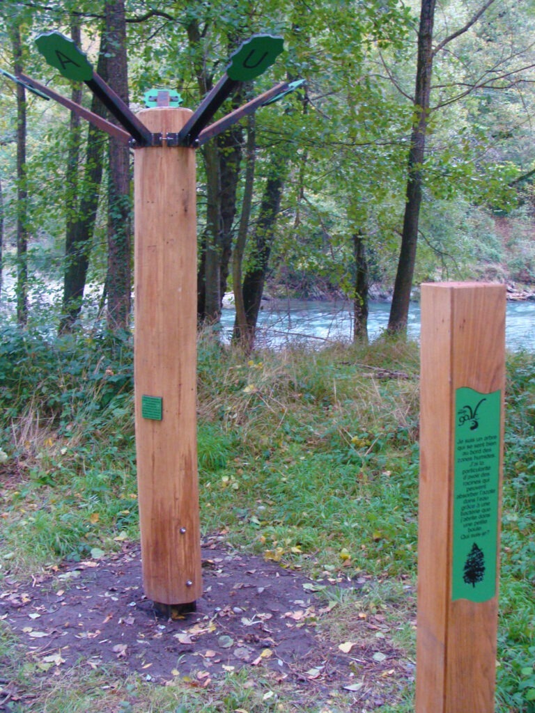 Photographie d'un "arbre" symbolique (tronc de bois usiné à 6 face et branche en métal) dont chaque branche porte une feuille sur laquelle est inscrite une lettre. L'ensemble des lettres forment un mot à découvrir. A l'avant-plan un mât en bois porte un petit cartel informatif associé au mot à découvrir. Concept et design Alexandre Drabzak.