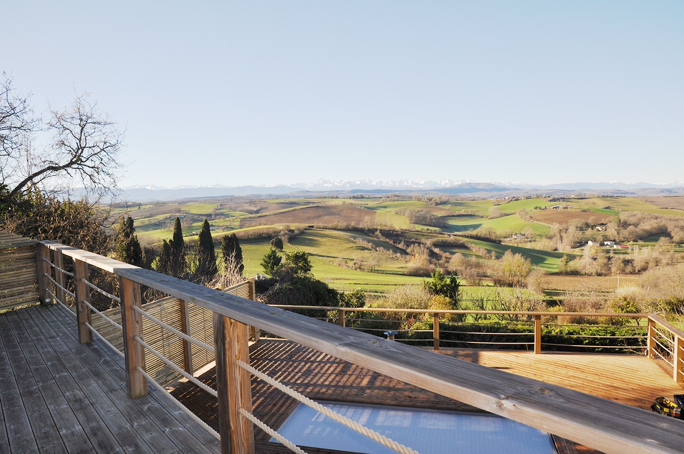 Point de vue sur les Pyrénées et le bocage ariégeois depuis la terrasse haute du jardin et son garde corps en bois et en corde. En avant plan :  grande terrasse en bois avec sa piscine.