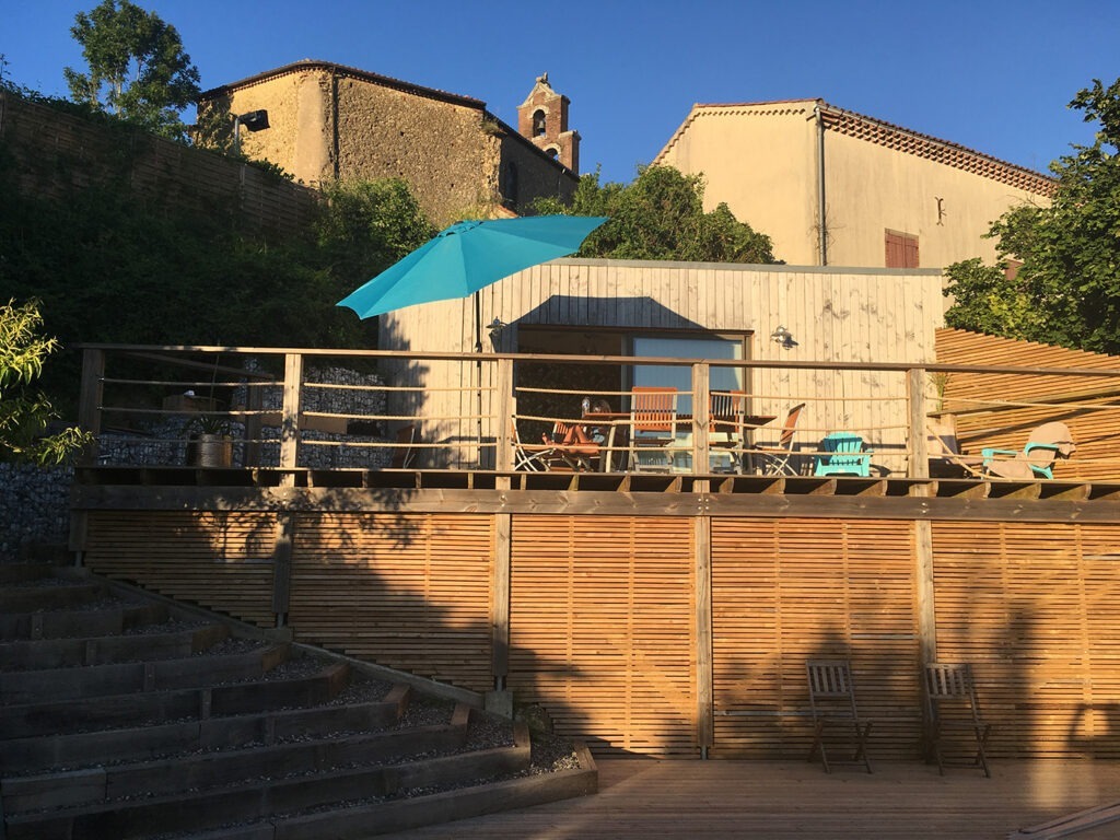 Photographie la terrasse supérieure du jardin, le pool-house et, en arrière-plan, l'église du village sur fond de ciel bleu. 