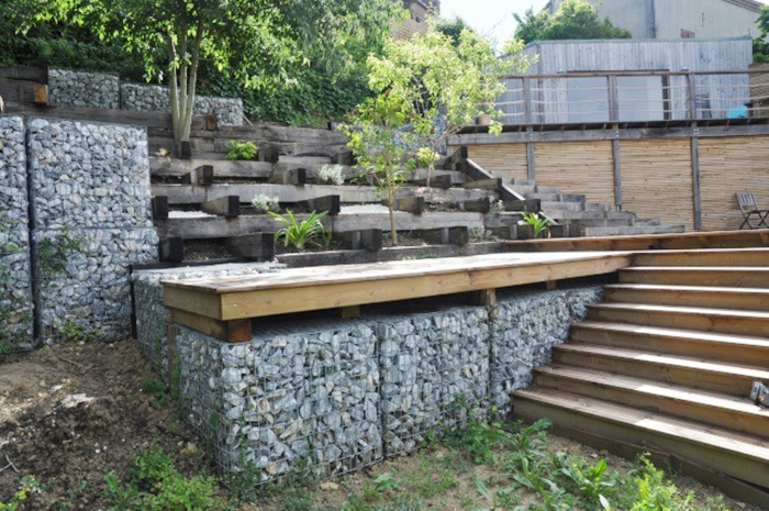 Photographie montrant un escalier et le dispositif de soutènement réalisé au moyen de gabions. Le plan intermédiaire montre un système de terrasses de culture réalisé en traverse de chêne.