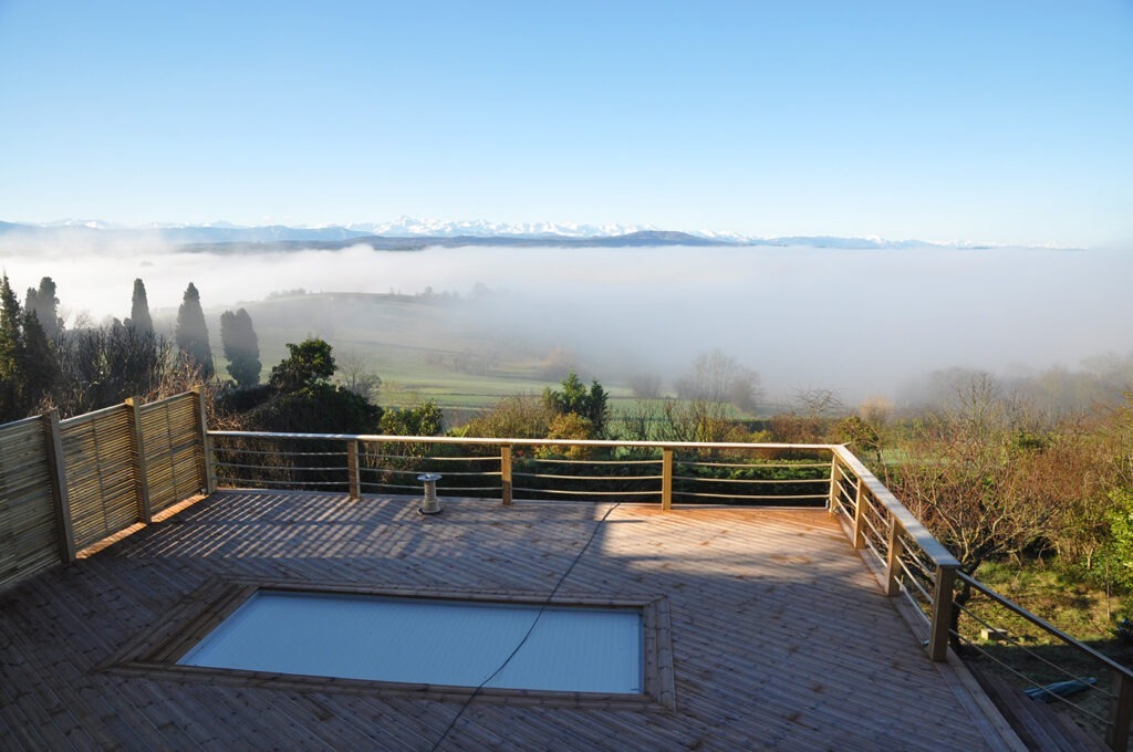 Photographie montrant le point de vue sur les Pyrénées et le paysage collinéen intermédiaire couvert d'une nappe de brouillard. Au premier plan la grande terrasse en bois, avec sa piscine en couloir de nage.