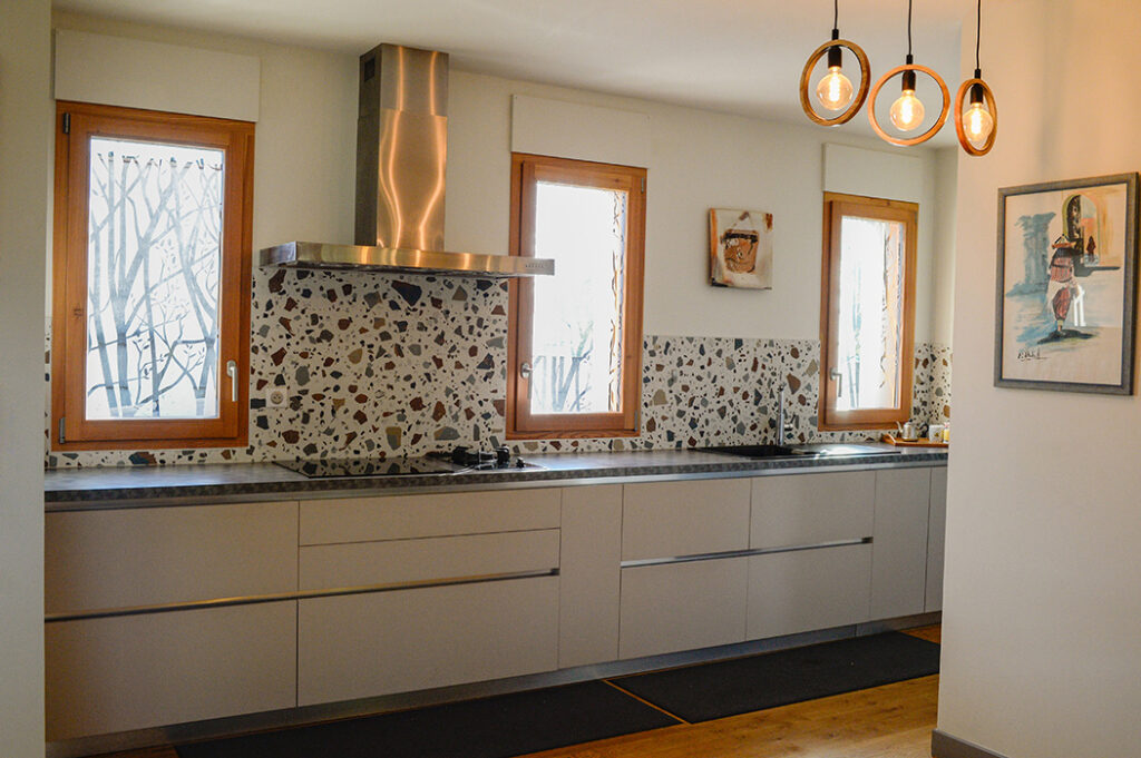 Photographie de la cuisine de style moderne (épurée), avec hotte en inox, crédence en faïence de style Terrazzo contemporain, luminaire et fenêtres en bois avec brise vue intégrée au vitrage.