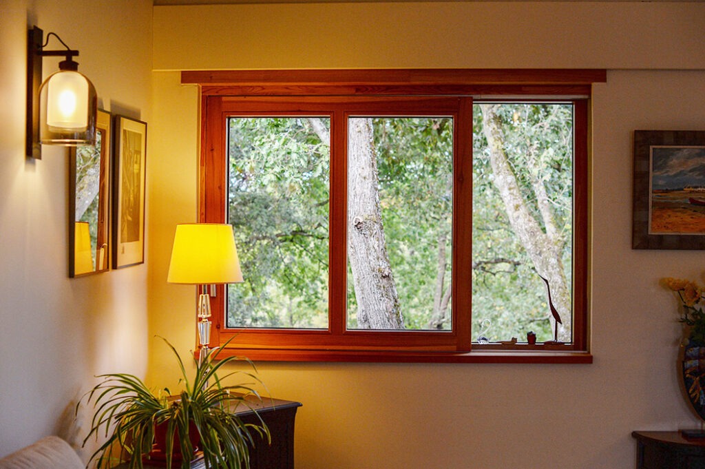 Photographie de la fenêtre coulissante en bois du salon donnant sur la canopée des arbres. Luminaires et décoration intérieure.