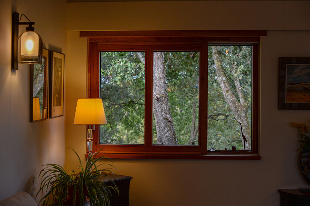 Photographie de la fenêtre coulissante en bois du salon donnant sur la canopée des arbres. Luminaires et décoration intérieure.