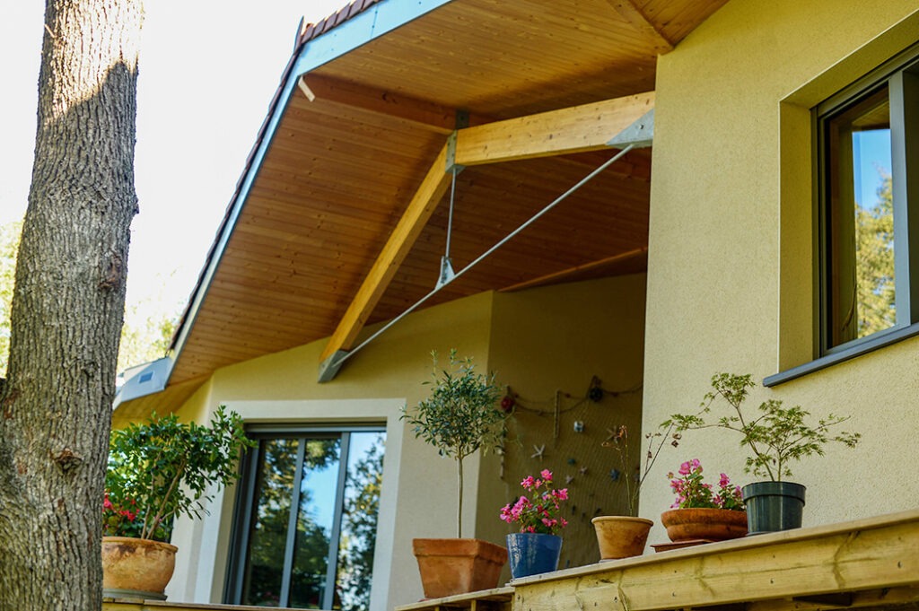 Photographie prise depuis l'extérieure montrant un auvent couvrant une alcôve, une charpente moderne mêlant bois et métal, une baie vitrée et une fenêtre en alu-bois et une terrasse en bois.