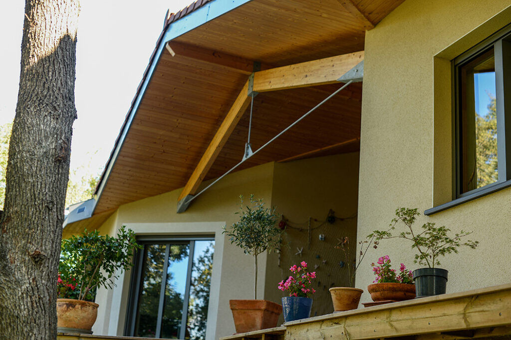 Photographie prise depuis l'extérieure montrant un auvent couvrant une alcôve, une charpente moderne mêlant bois et métal, une baie vitrée et une fenêtre en alu-bois et une terrasse en bois.