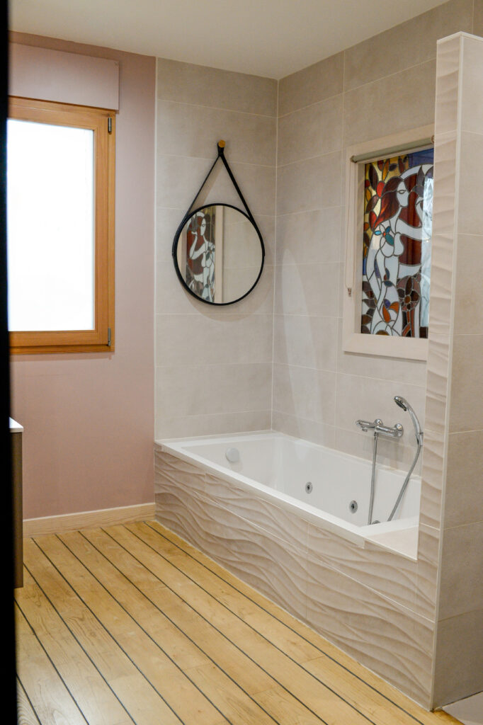 Photographie de la salle de bain avec plancher bois "pont de bateau", baignoire, vitrail, faïence, miroir et fenêtre à vitrage opalescent.