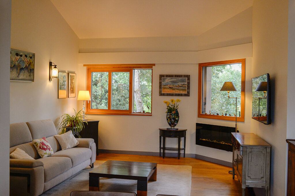 Photographie du salon meublé, avec plafond rampant, luminaires et fenêtres donnant sur la canopée des arbres.