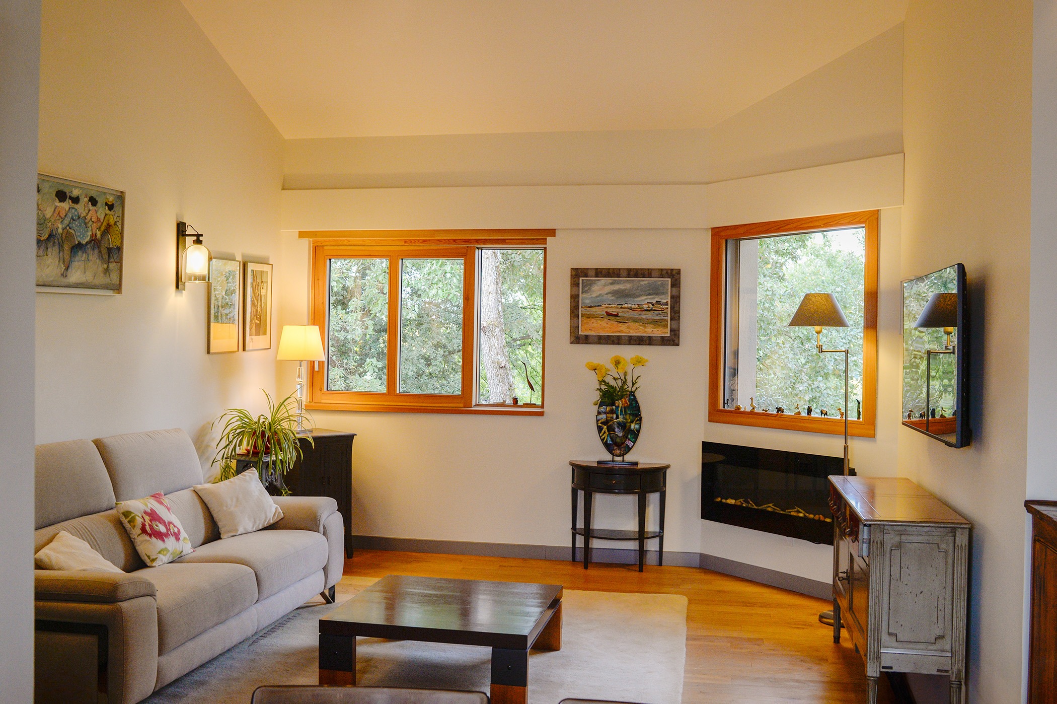 Photographie d'un salon meublé, avec plafond rampant, luminaires et fenêtres donnant sur la canopée des arbres. 