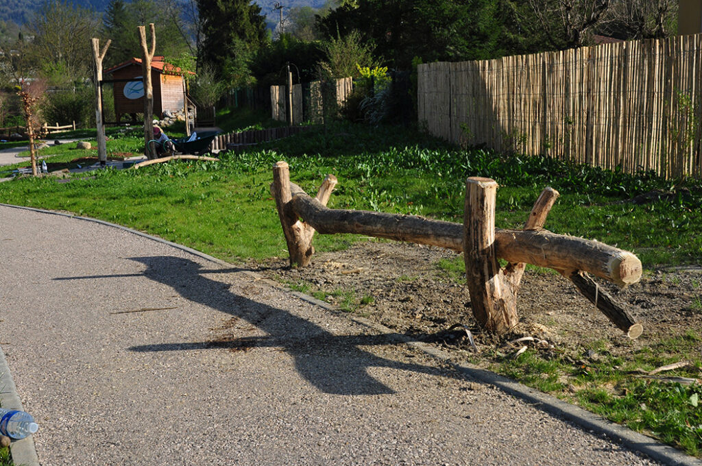 Banc en bois "sauvage", fait de troncs de bois brut écorcé. Le dispositif est un tronc principal horizontal encastré dans deux tronc formant des fourches et enterrés dans le sol. L'ensemble est installé au sein d'un espace pédagogique dédié à la vie des rivières et la pêche.