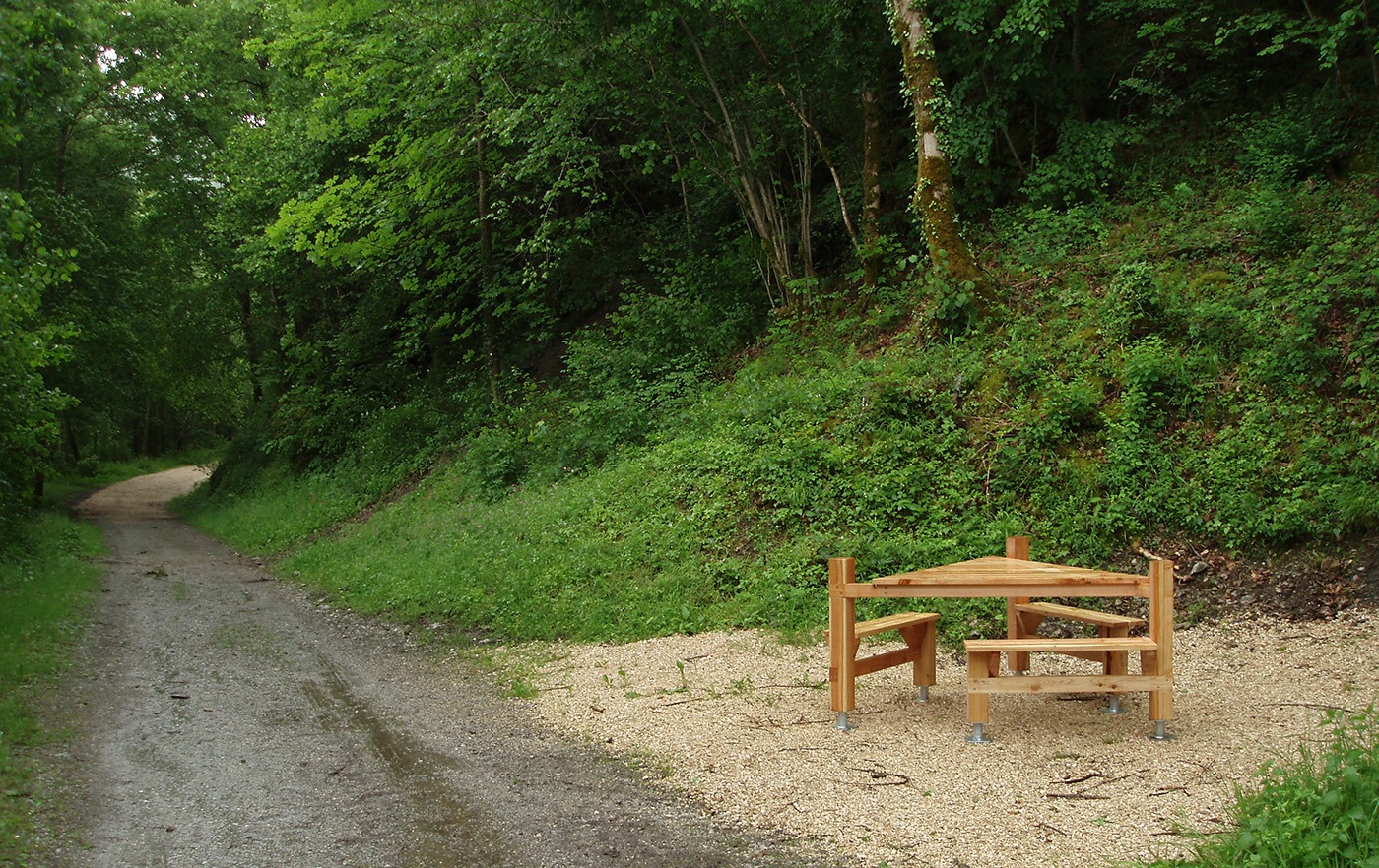Table de pique-nique design en bois en forme de triangle et installée au bord d'un chemin dans un espace naturel verdoyant.
