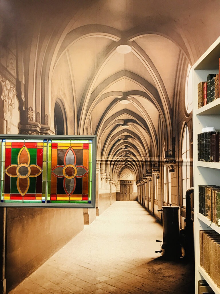 Photographie en perspective du musée montrant une collection de livres anciens dans une bibliothèque, la grande photographie de l'ancien cloître et le vitrail découvert lors des travaux.