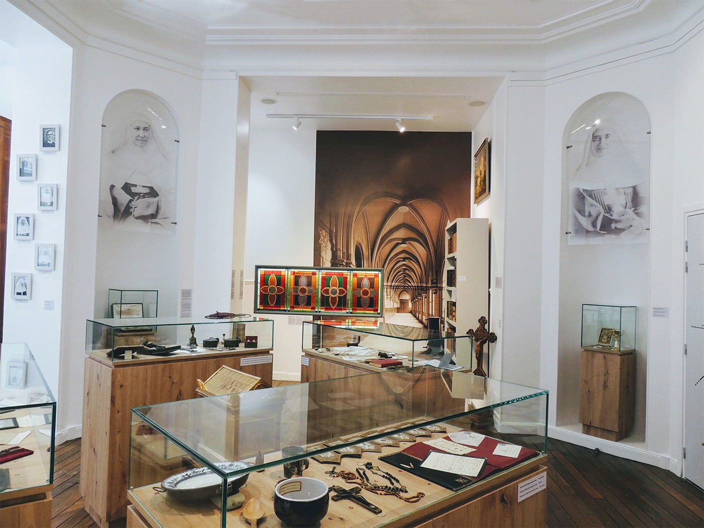 Photographie du musée montrant plusieurs vitrines contenant des objets historiques, les grands portraits de Mère Marie-Eugénie et Mères Thérèse-Emmanuel, fondatrices de la congrégation, et, en fond, la grande photographie de l'ancien cloître et le vitrail.