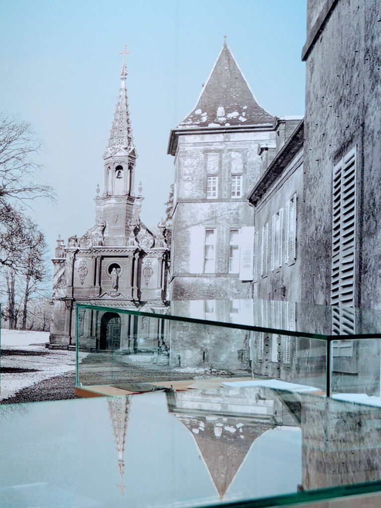 Prise de vue artistique de la grande photographie du Château de Preisch en Lorraine, dont la chapelle de Baptême de Marie-Eugénie, représentée en noir et blanc avec effet de colorisation (ciel bleu), se reflétant sur le verre des vitrines.