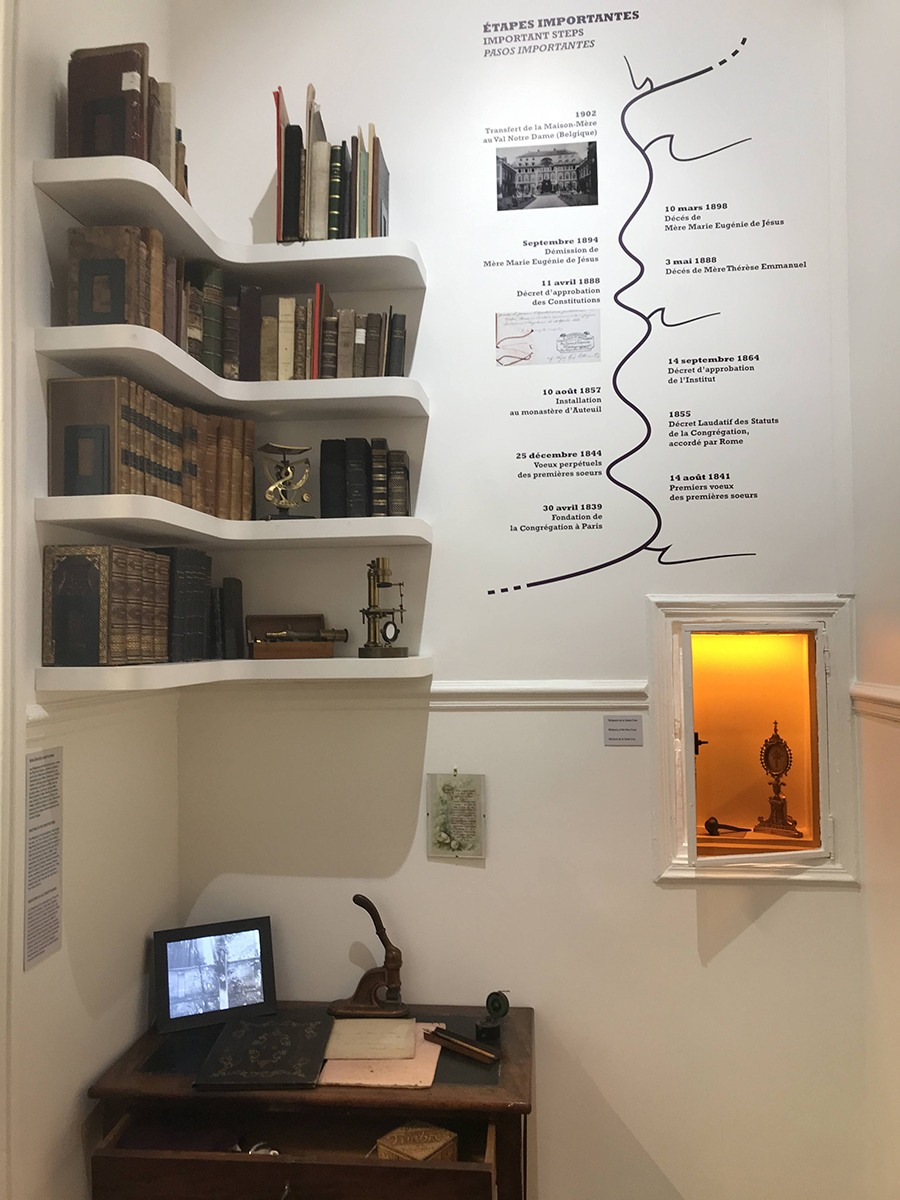 Petit bureau de Mère Marie-Eugénie, fondatrice des Soeurs de l'Assomption, étagère moderne avec livres anciens, la quatrième partie de la frise chronologique, portant sur les évènements marquant la reconnaissance de la congrégation, et niche de crédence comprenant des objets liturgiques.