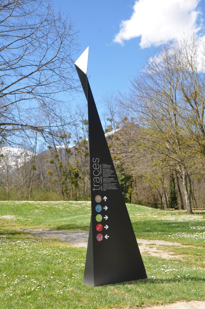 Photographie d'un totem design, en acier plié et thermolaqué, en cohérence avec l'architecture contemporaine marquée du site. Le principe design n'est pas sans rappeler les origamis. Celui-ci, destiné à marquer et informer un lieu d'exposition ou d'animation extérieur, comprend le nom et la description de l'atelier, ainsi que des symboles directionnels.