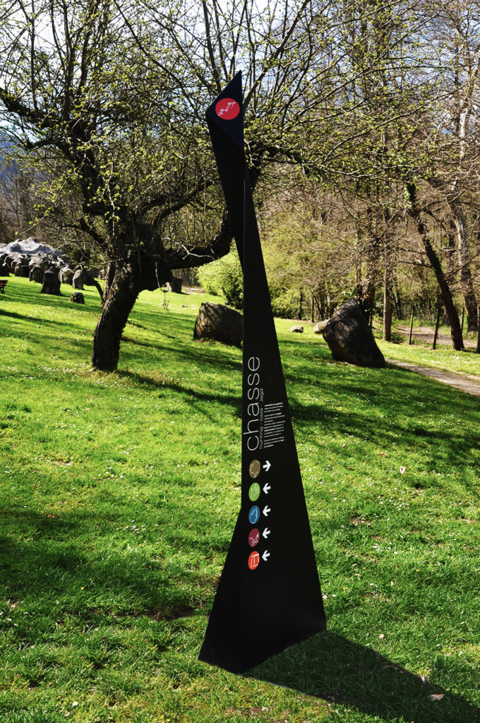 Photographie d'un totem design, en acier plié et thermolaqué, en cohérence avec l'architecture contemporaine marquée du site. Le principe design n'est pas sans rappeler les origamis. Celui-ci, destiné à marquer et informer un lieu d'exposition ou d'animation extérieur, comprend le nom et la description de l'atelier, ainsi que des symboles directionnels.