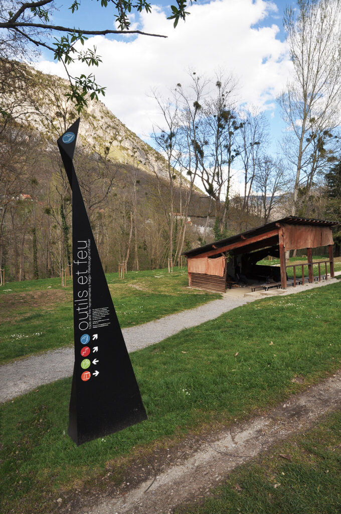 Photographie d'un totem design, en acier plié et thermolaqué, en cohérence avec l'architecture contemporaine marquée du site. Le principe design n'est pas sans rappeler les origamis. Celui-ci, destiné à marquer et informer un lieu d'exposition ou d'animation extérieur, comprend le nom et la description de l'atelier, ainsi que des symboles directionnels.