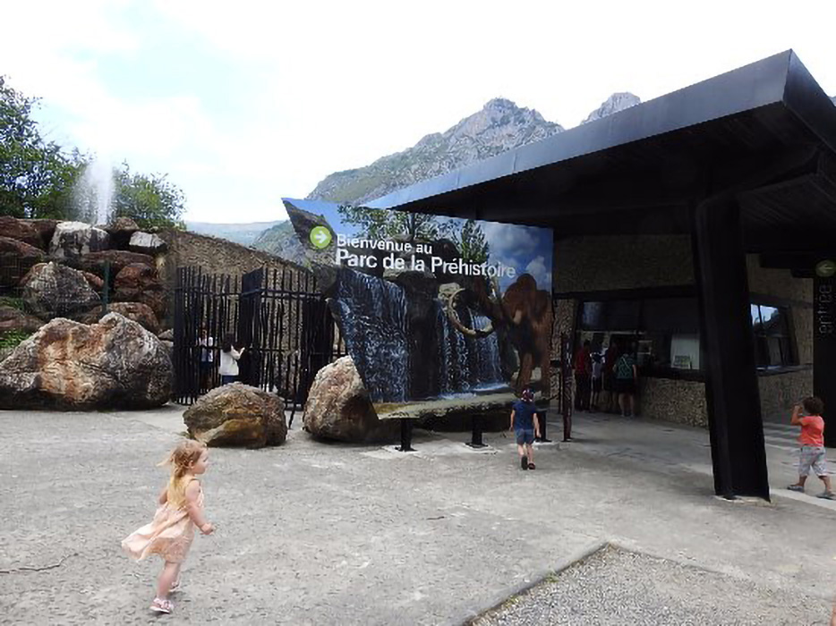 Photographie en grand angle du bâtiment du Parc de la préhistoire vu depuis l'accès du parking. La prise de vue montre les lignes et couleurs contemporaines du bâtiment, son guichet d'accueil, le grand panneau marquant l'entrée, et une famille de visiteurs.
