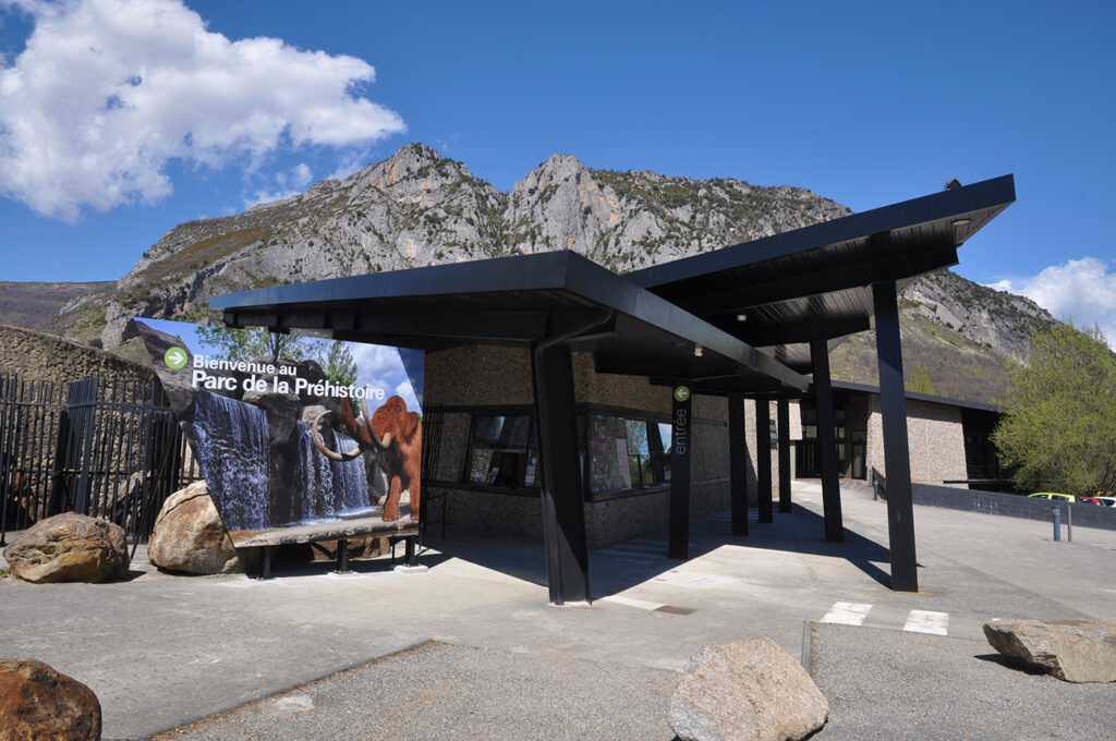 Photographie en grand angle du bâtiment du Parc de la préhistoire vu depuis l'accès du parking. La prise de vue montre les lignes et couleurs contemporaines du bâtiment, son guichet d'accueil et le grand panneau marquant l'entrée.
