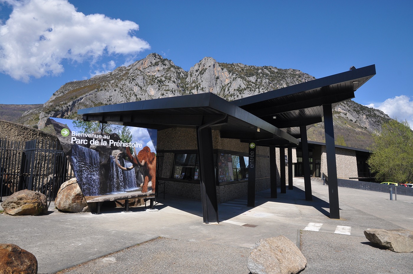 Photographie en grand angle du bâtiment du Parc de la préhistoire vu depuis l'accès du parking. La prise de vue montre les lignes et couleurs contemporaines du bâtiment, son guichet d'accueil et le grand panneau marquant l'entrée.