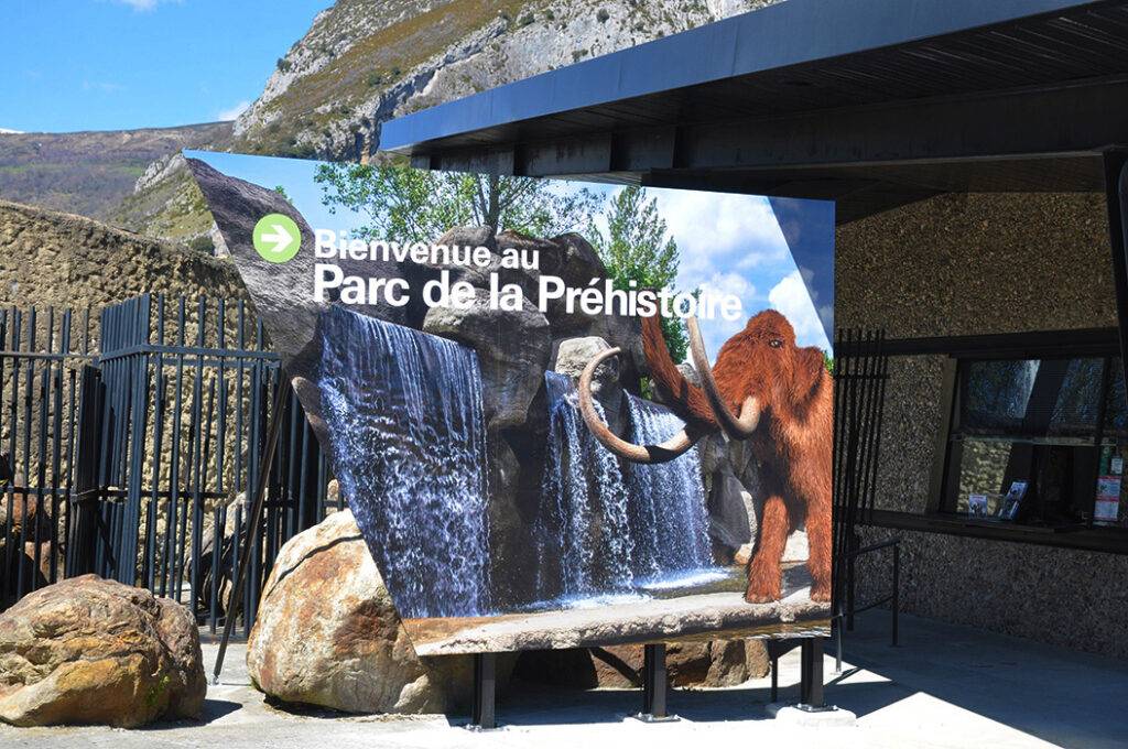 Photographie du Grand panneau marquant l'entrée du site. Son design reprend l'esprit des lignes du bâtiment existant, visible en arrière plan sur la prise de vue, dont le guichet d'accuiel.