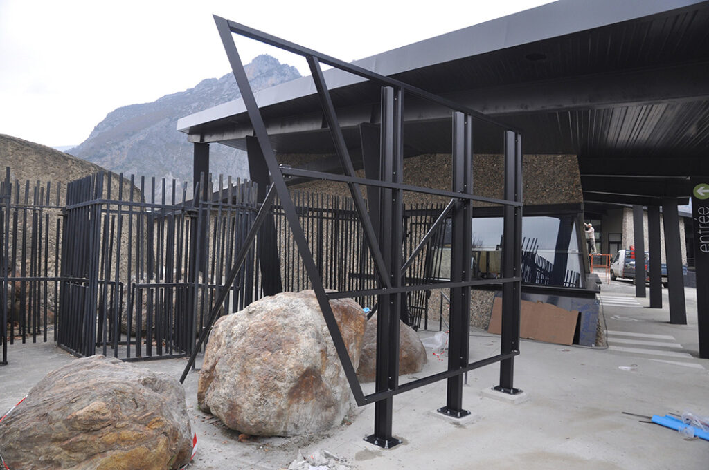 Photographie de la structure en métal thermolaqué noir, destiné à recevoir le Grand panneau marquant l'entrée du site. Son design reprend l'esprit des lignes du bâtiment existant, visible en arrière plan sur la prise de vue.