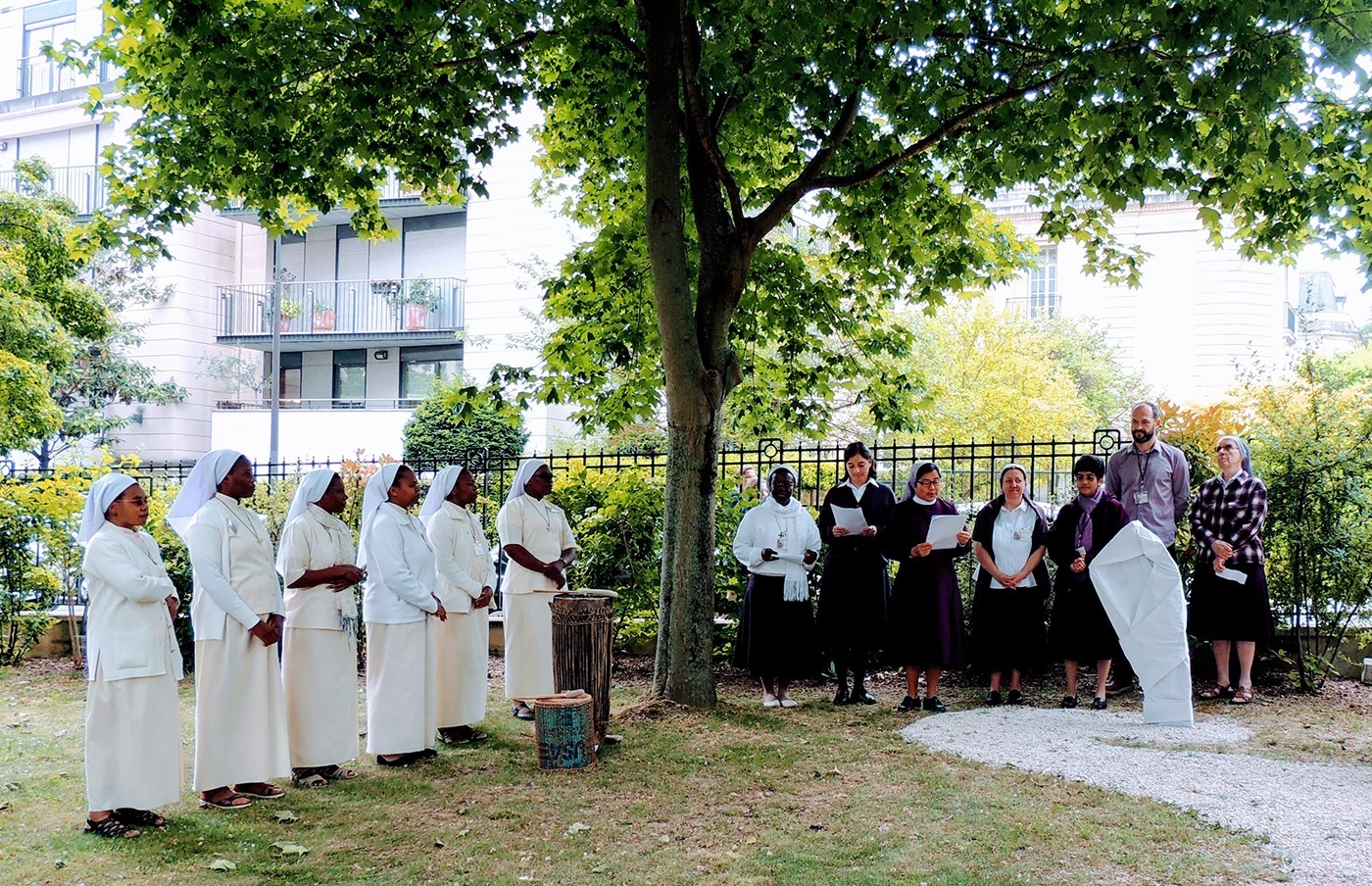 Photographie de la cérémonie d'inauguration du parcours historique précédant la présentation au public du lutrin design (masqué par un tissu blanc). Visible sur la photographie, à gauche un groupe de Sœurs africaines vêtues de blanc animant la cérémonie de chants et de musiques traditionnelles, et à droite un autre groupe de Sœurs lisant des textes, dont Mère Rékha et Sœur Véronique (co-conceptrice du parcours), au milieu desquelles se trouve Alexandre Drabzak (designer du Lutrin et co-concepteur du parcours),