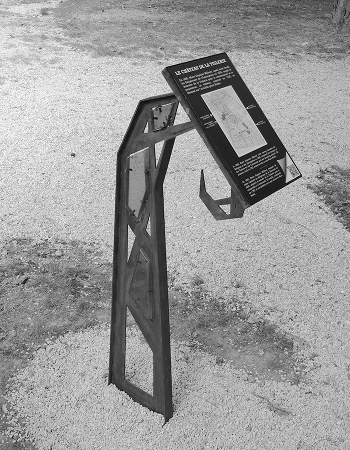 Photographie d'un lutrin design installé dans le parc du Couvent des Soeurs de l'Assomption. Le lutrin est vu de trois-quart face, comporte un Cartel sur le "Château de la Tuilerie" et présente des vitraux bleu, vert et rouge.