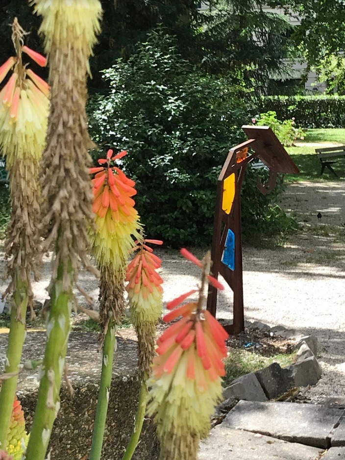 Photographie d'un lutrin installé proche de la chapelle (non visible) avec fleurs de Kniphofia d'un jaune et orange presque fluorescents en premier plan. Le lutrin est vu de dos et présente des vitraux bleu, jaune opaque et orange.