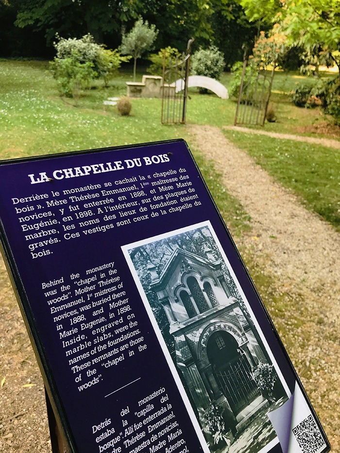 Photographie en perspective du cartel d'un des lutrins du parcours historique, dédié à la chapelle du bois aujourd'hui disparu. A l'arrière plan, les vestiges conservés sur place de l'ancienne chapelle.