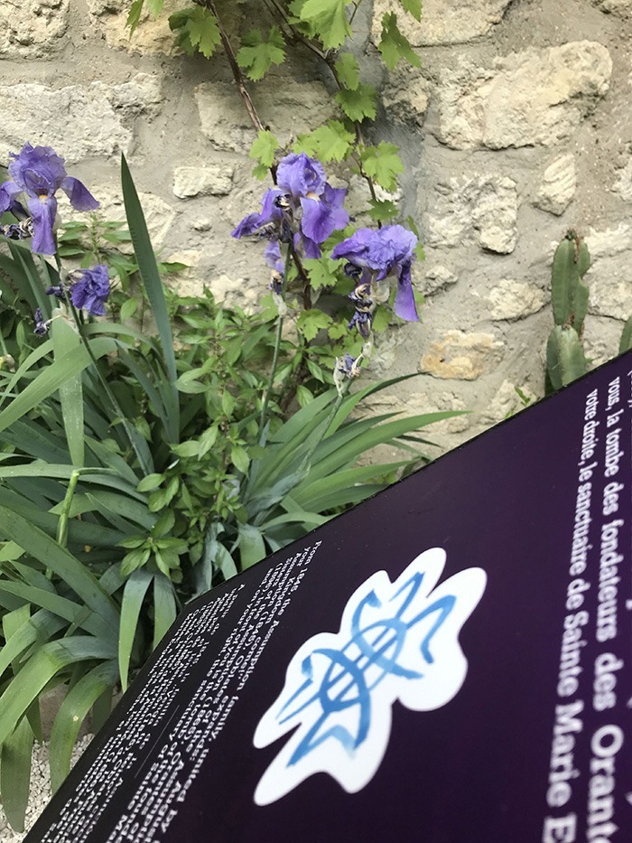 Photographie en perspective du cartel d'un des lutrins du parcours historique, dédié à la famille assomption. A l'arrière plan, un mur de pierre et des iris violet.
