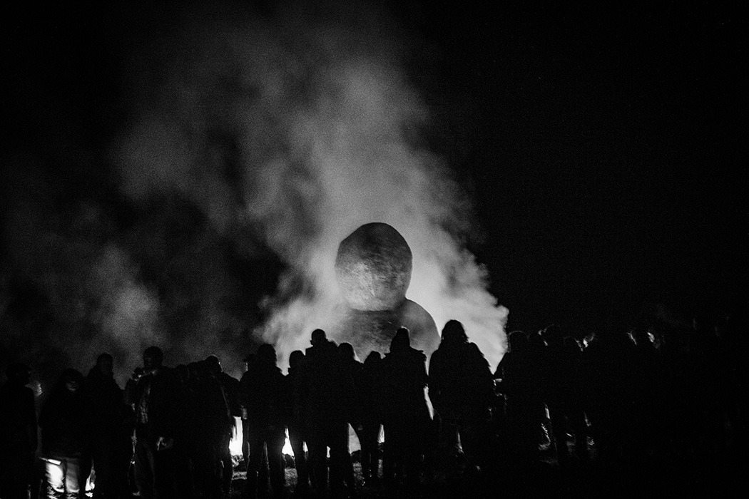 Photographie en noir et blanc montrant les silhouettes du public autour du cercle de feu entourant la Vénus monumentale.