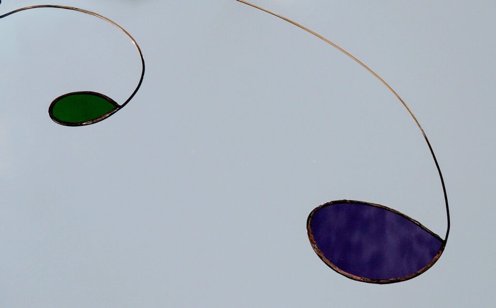 Photographie rapprochée d'un mobile montrant un vitrail bleu ciel découpé en goutte d'eau et serti par une tige de laiton qui prolonge la forme.