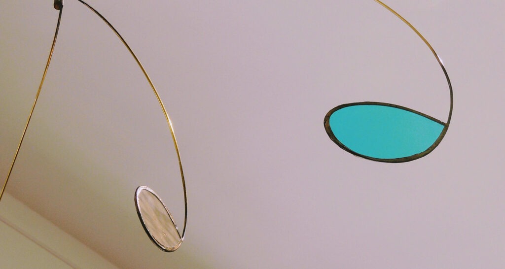 Photographie rapprochée d'un mobile montrant un vitrail bleu ciel découpé en goutte d'eau et serti par une tige de laiton qui prolonge la forme.