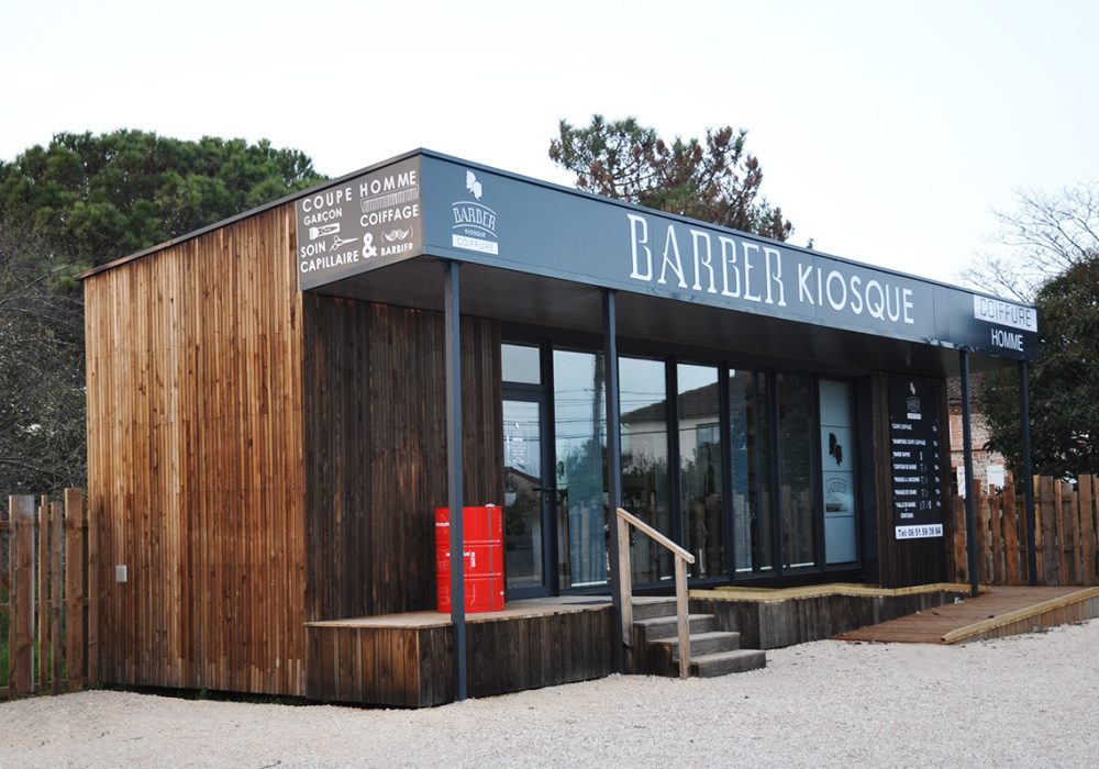 Salon de coiffure-Barber créé à partir d'un conteneur maritime. Bardage en bois brut grisé, coursive couverte et grande vitrine constitue une apparence modernisée de "saloon" de western.
