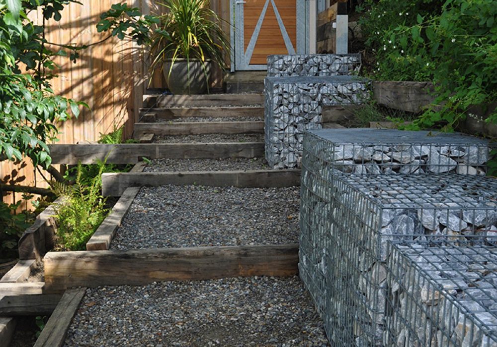 Larges marches de jardin structurées en traverses paysagères en chêne, associées à des gabions. La photographie montre la descente depuis la porte d'entrée design - en bois et métal - du jardin. La construction appartient à un grand jardin, initialement très pentu et aménagé en terrasses, afin de valoriser le point de vue sur les Pyrénées.